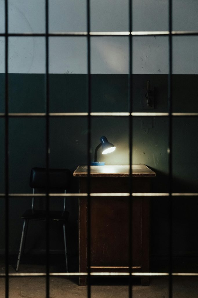 A dimly lit prison cell with a lamp on a table behind bars.