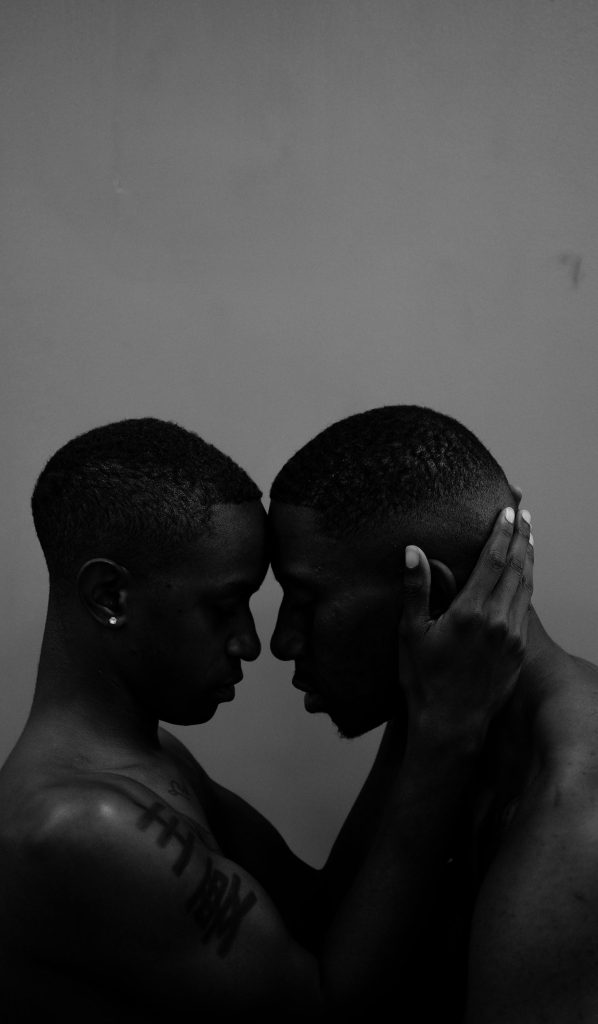 Two shirtless men share a tender moment, embodying love and connection in a black and white setting.