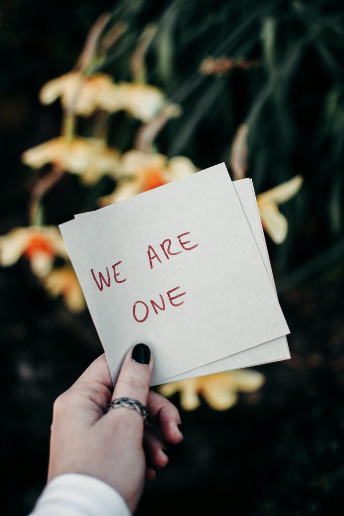 A hand holding a note reading 'We Are One' against a floral outdoor backdrop.